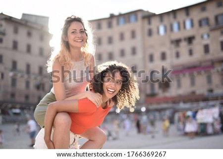 A blonde girl on the back of a black-haired lady behind buildings showing happiness and excitement