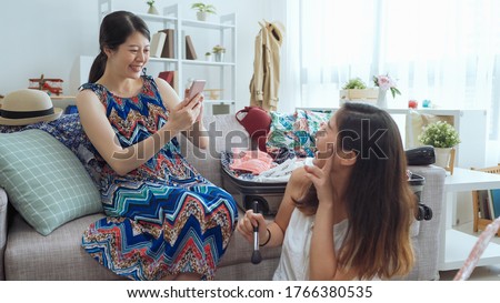 young girl in fashion summer style dress sitting on couch and holding mobile phone taking picture of her friend. female roommate turn around holding brush and showing victory sign looking cellphone