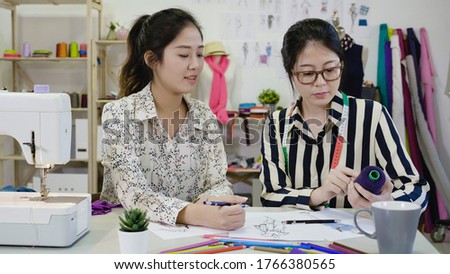 Two young professional fashion designer colleagues women working on project of new collection. coworkers dressmakers drawing sketches for clothes in workplace and choosing color of thread at table.