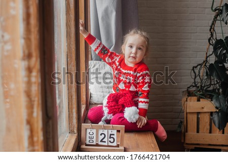 Little cute girl in a New Year hat and a sweater sits at the window.
Merry christmas and happy New Year
