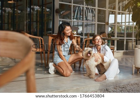 Summer. Women With Pet On Terrace In Dog-Friendly Cafe. Fashion Girls In Casual Outfit With Puppy Sitting On Patio And Looking Away. Vacation With Dog As Part Of Modern Lifestyle. 