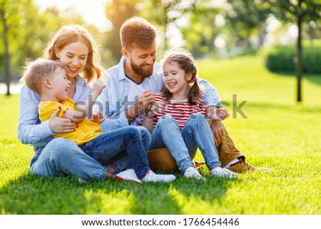 Cheerful parents smiling and playing with kids while sitting on green grass on sunny summer day in park
