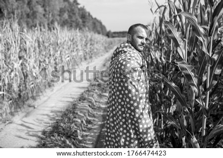 Black and white picture of beautiful caucasian man with short dark hair in wrapped in big red and white coverlet walks to the big cornfield relaxes and thinks about something.