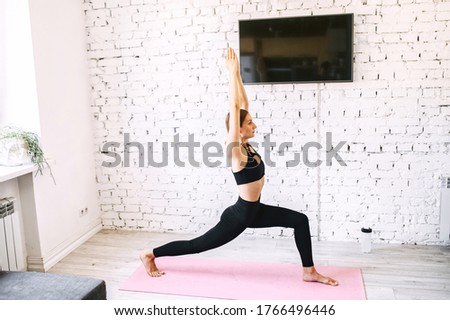 Side long shot of a young girl practicing yoga. She stands on a mat with her legs bent in knees, hands are put together in the air. She is in her sportswear standing against white brick wall