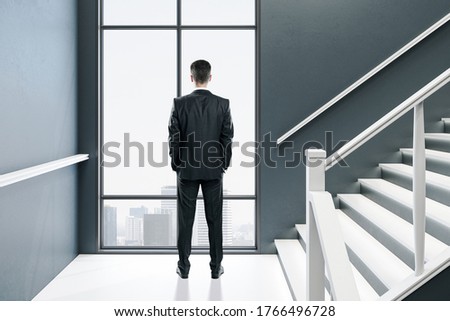Businessman looking in window in modern concrete hall interior with stairs and city view. Art and design concept. Mock up,