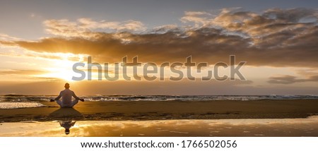 yoga meditation silhouette lotus sunrise beach, mindfulness, wellness and wellbeing concept, water reflection of man in yoga lotus pose sitting alone on sand with ocean cloud background, copy space Royalty-Free Stock Photo #1766502056
