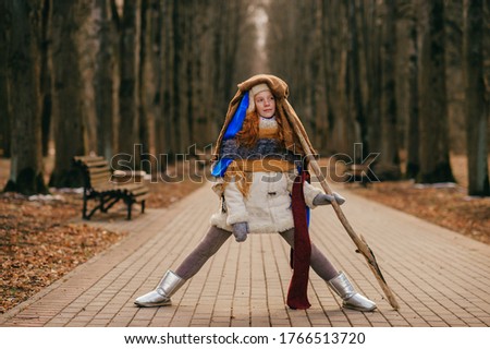 Picture of beautiful girl in warm clothes wrapped in several scarves with long wooden stick walking through the autumn park.