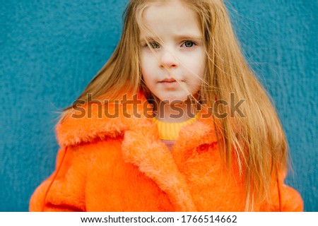 A beautiful little girl with medium blonde hair in a bright orange fur coat looks at the camera, picture isolated on blue background