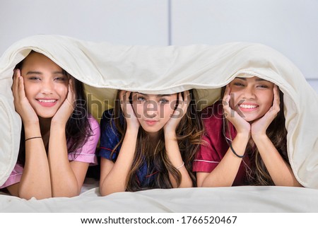 Group of happy teenage girls looking at the camera on the bed while lying under blanket in bedroom at home