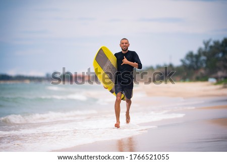 Fit surfer man running with yellow surfboard longboard on the sunny beach showing a Hawaiian shaka sign. Summer vacation active hobby.