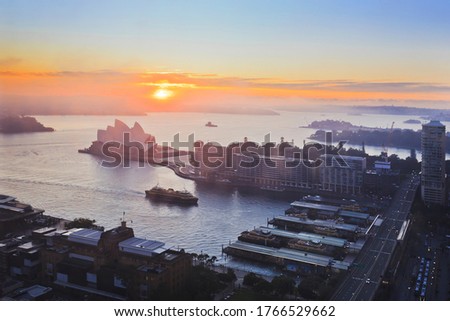 Bright rising sun over Sydney harbour around major Sydney city landmarks on waterfront with ferry wharves in Circular Quay.