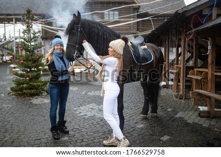 Picture of two young attractive sisters in warm jackets, trousers, boots, scarfs and knitted hats with a horse outside in winter