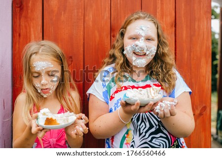 Picture of cute caucasian girl with long fair hair and in pink dress with her older chubby sister smiles and eat a cacke against the wooden wall in village