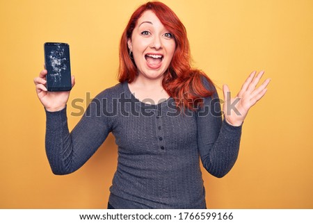 Young beautiful redhead woman holding broken smartphone showing cracked screen celebrating achievement with happy smile and winner expression with raised hand