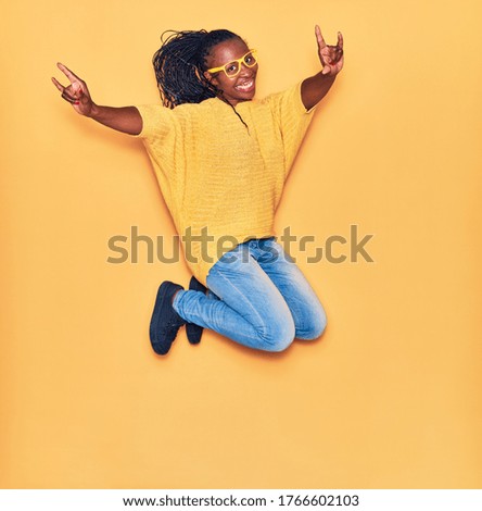 Young beautiful african american woman wearing casual clothes and glasses smiling happy. Jumping with smile on face doing horns sign over isolated yellow background