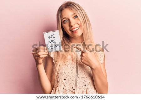 Young beautiful blonde girl holding we are equal paper smiling happy pointing with hand and finger 