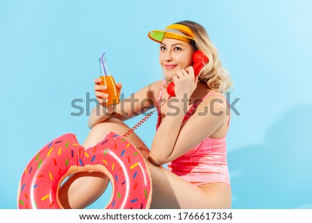 portrait of happy young woman in bathing suit sitting with rubber ring, cocktail and talking on phone handset and looking away with smile. indoor studio shot isolated on blue background