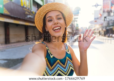 Young brunette woman using a mobile phone takes a selfie with attractive smile. Happy Hispanic girl traveler wear summer dress with hat waving to internet audience. Influencer and vlogging concept