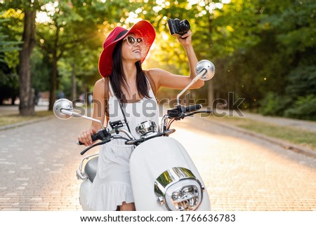 attractive smiling woman riding on motorbike in street in summer style outfit wearing white dress and red hat traveling on vacation, taking pictures on vintage photo camera