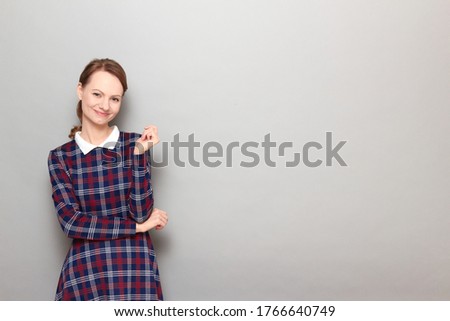 Studio portrait of pretty cheerful blond girl wearing checkered dress, smiling joyfully, looking relaxed and successful, with positive facial emotion, standing over gray background, with copy space