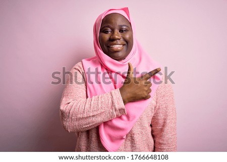 Young african american plus size woman wearing muslim hijab over isolated pink background cheerful with a smile of face pointing with hand and finger up to the side with happy and natural expression