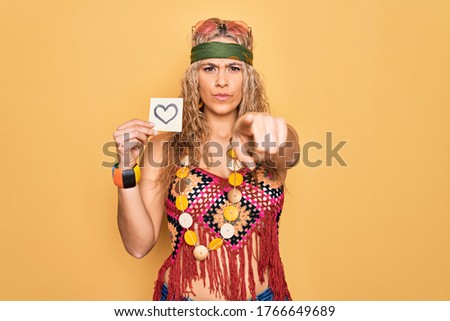 Beautiful blonde hippie woman wearing sunglasses and accessories holding reminder with heart pointing with finger to the camera and to you, confident gesture looking serious