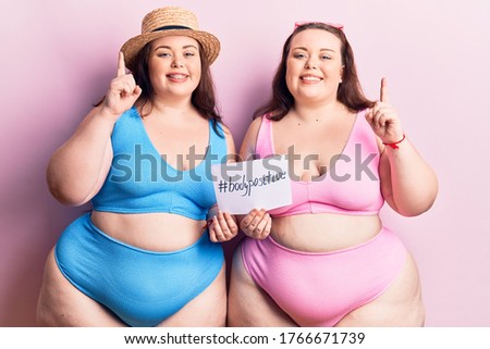 Young plus size twins wearing bikini holding paper with hashtag body positive smiling with an idea or question pointing finger with happy face, number one 