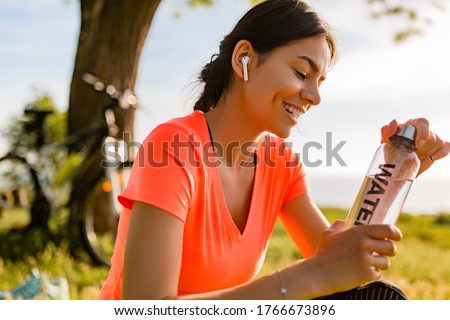 smiling beautiful woman drinking water in bottle doing sports in morning in park nature doing yoga on mat, pink fitness outfit, happy healthy lifestyle, music in earphones, bicycle on background