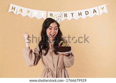 Image of excited woman making winner gesture while posing with birthday cake isolated over beige background