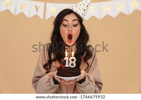 Image of young woman in party cone blowing out candles on birthday cake isolated over beige background