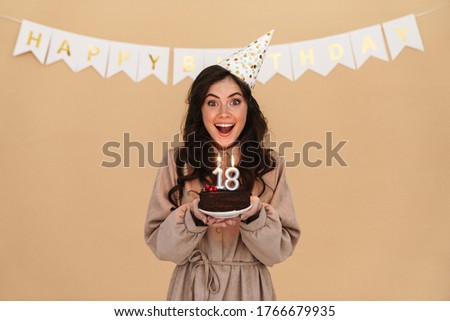 Image of excited young woman in party cone smiling while posing with birthday cake isolated over beige background