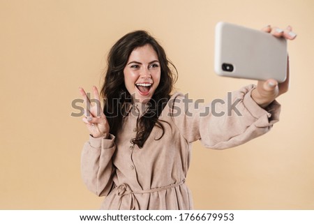 Image of joyful woman gesturing peace sign while taking selfie on cellphone isolated over beige background