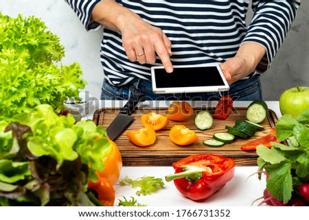 Woman cooking and holding tablet computer with blank screen in hand. Cutting vegetable ingredients and following recipe from the internet.