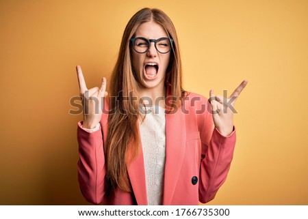 Young beautiful redhead woman wearing jacket and glasses over isolated yellow background shouting with crazy expression doing rock symbol with hands up. Music star. Heavy concept.