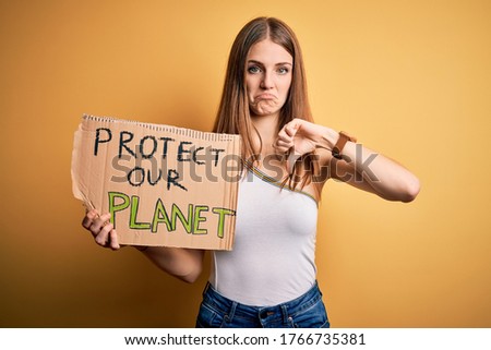 Young redhead woman asking for enviroment holding banner with protect planet message with angry face, negative sign showing dislike with thumbs down, rejection concept