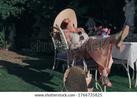Attractive and young adult woman enjoying summer vacation in her home garden with elegant accessories and accessories such as hat, sunglasses and handbag with a very chic style