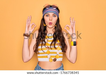 Young beautiful hippie woman with blue eyes wearing accesories and sunnglasses looking surprised and shocked doing ok approval symbol with fingers. Crazy expression