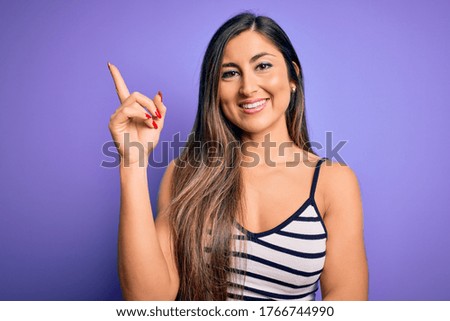 Young beautiful woman wearing casual summer striped top over purple isolated background with a big smile on face, pointing with hand and finger to the side looking at the camera.