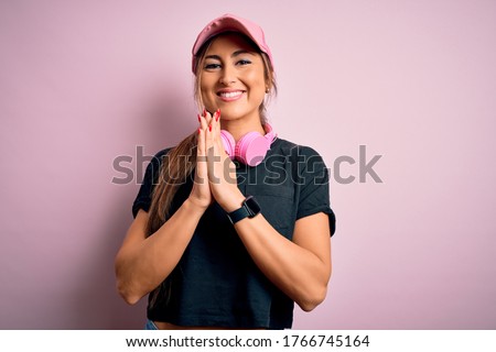 Young beautiful fitness sports woman wearing training cap and headphones over pink background praying with hands together asking for forgiveness smiling confident.