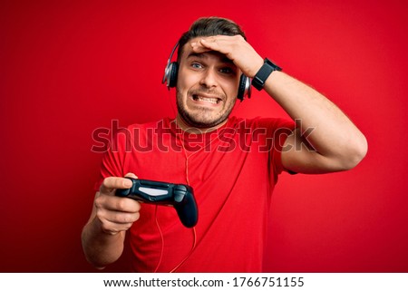 Young gamer man with blue eyes playing video games using gamepad joystick stressed with hand on head, shocked with shame and surprise face, angry and frustrated. Fear and upset for mistake.