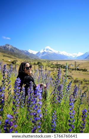Picture of a smiling woman wearing sunglasses sitting amongst purple lupin wild flowers in the grassy meadow with snow capped mountain peaks of Mount Cook in the background