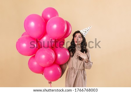 Image of surprised cute woman in party cone posing with pink balloons isolated over beige background