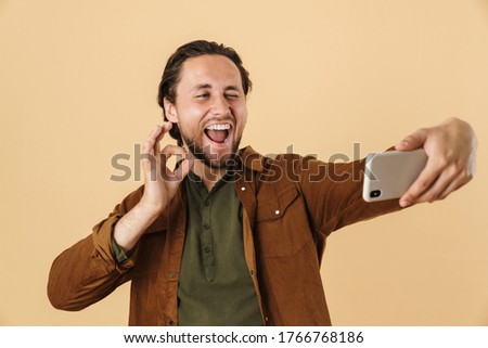 Image of delighted man showing ok sign while taking selfie on cellphone isolated over beige background