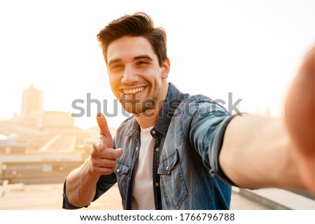 Photo of joyful unshaven young man pointing finger at camera while taking selfie photo outdoors