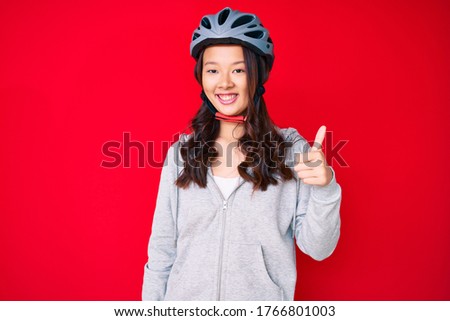 Young beautiful chinese girl wearing bike helmet smiling happy and positive, thumb up doing excellent and approval sign 
