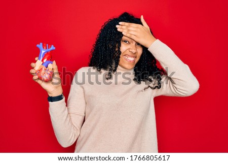 Young african american curly woman holding plastic heart standing over red background stressed with hand on head, shocked with shame and surprise face, angry and frustrated. Fear and upset for mistake