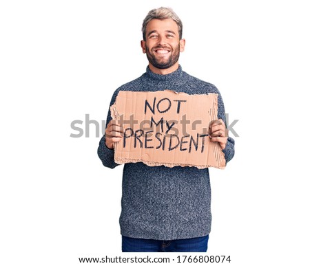 Young handsome blond man holding not my president cardboard banner looking positive and happy standing and smiling with a confident smile showing teeth 