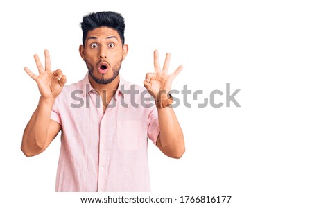 Handsome latin american young man wearing casual summer shirt looking surprised and shocked doing ok approval symbol with fingers. crazy expression 