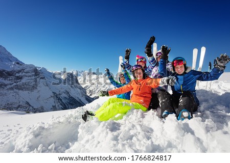 Group of kids in colorful ski sport snow outfit rise hands sitting together over mountain tops