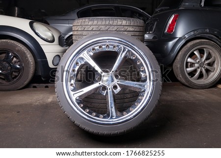 Automobile composition made up stack of tires and wheel with shiny metal disc in the foreground against the background of cars in a car service before seasonal replacement or after breaking through. Royalty-Free Stock Photo #1766825255